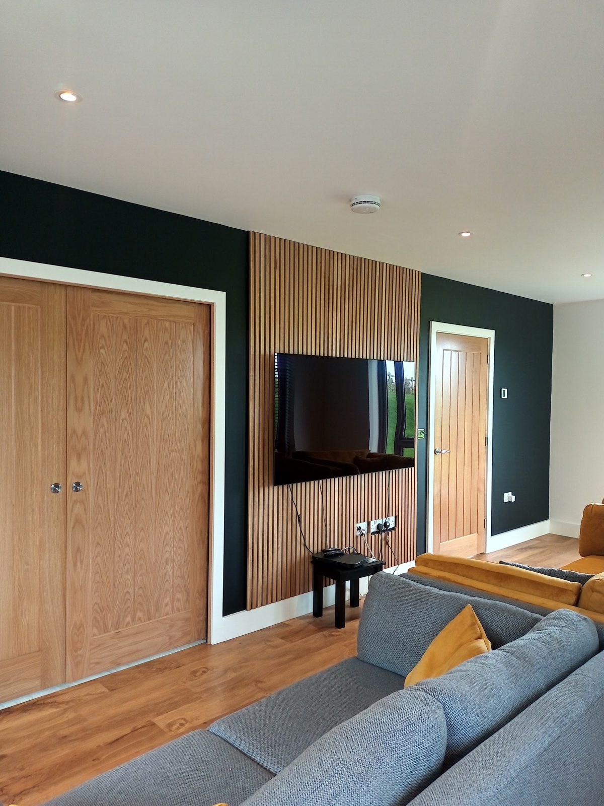 Living room with deep green walls, a timber slat panel behind the TV and oak doors